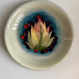 May include: A white ceramic dish with a blue and white floral design. The dish features a stylized flower with red, yellow, and green leaves. The flower is surrounded by a ring of dark blue and red berries.
