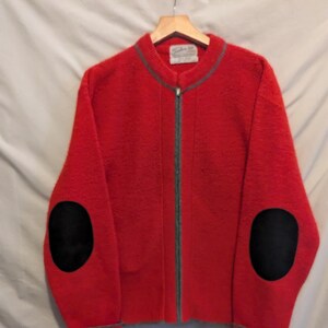 May include: Red zip-up cardigan with black elbow patches and grey trim. The cardigan has a pocket on the left side and a label that reads "Schuler Knit".