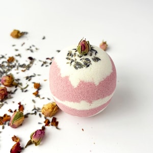 May include: A round bath bomb with a pink and white striped design, topped with a rose bud and dried lavender. Scattered around are dried rose buds and petals. The bath bomb is a self-care product.