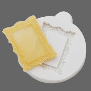 May include: White silicone mould for making a picture frame cake decoration. The mould has a detailed ornate design. A yellow picture frame made from the mould is shown next to it.