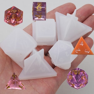 Puede incluir: Un conjunto de seis moldes de silicona transparentes para hacer dados. Los moldes son de varias formas, incluyendo un cubo, una pirámide y un dodecaedro. También hay varios dados rosas y naranjas en la imagen.