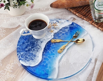 May include: A round blue and white resin tray with a wave design. The tray has a white rim and features gold-colored silverware and seashells.
