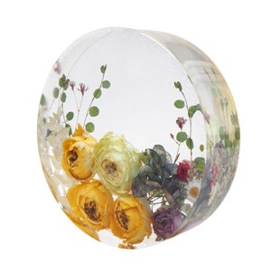 May include: A clear, round resin paperweight with dried flowers. The flowers include yellow roses, purple and white blooms, and green foliage. The paperweight is a decorative item for a desk or shelf.