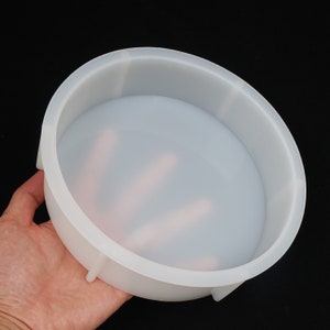 以下が含まれることがあります： 浅いボウルの形をした、透明な丸いシリコンモールド。 モールドは滑らかな表面を持ち、黒い背景に対して手で保持されています。 モールドには小さな足と隆起したエッジがあります。