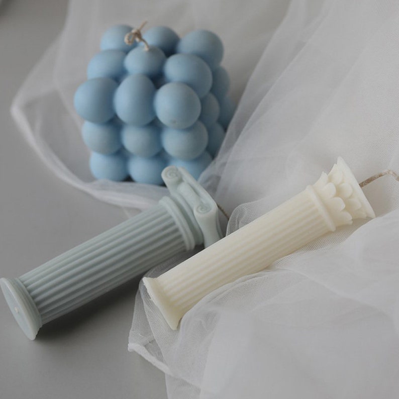 May include: Two decorative pillar candles, one light blue and one white, with intricate details. A blue bubble candle is also in the image.