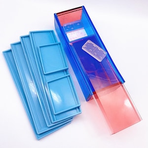 May include: A set of blue plastic domino tile holders with a clear plastic domino tile inside. The holders are stacked on top of each other. The domino tile is on top of a red plastic holder.