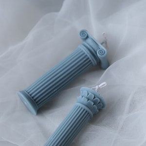 May include: Two blue pillar candles with intricate details on the top. The candles are on a white fabric background.