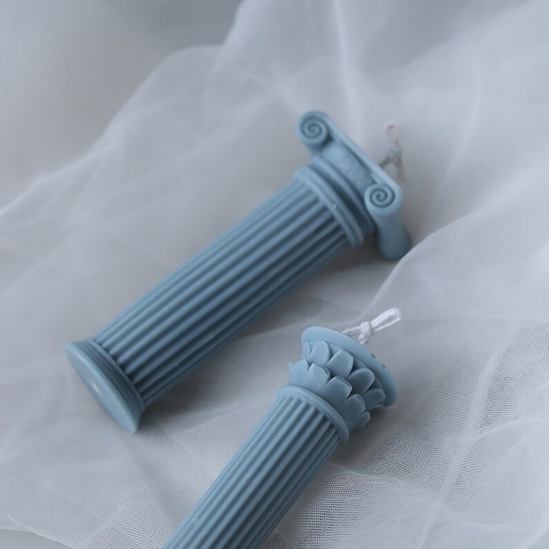May include: Two blue pillar candles with intricate details on the top. The candles are on a white fabric background.