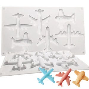 Peut inclure: Moule en silicone blanc avec 9 formes d'avions. Le moule est ouvert et montre le dessous avec 9 formes d'avions plus petites. Trois bonbons en forme d'avion finis sont présentés en bleu, rose et jaune.
