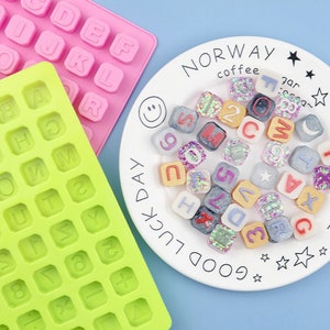 Puede incluir: Dos moldes de silicona para hacer formas de letras del alfabeto. Un molde es verde y el otro es rosa. Un plato blanco con el texto "NORWAY coffee" y "GOOD LUCK DAY" contiene varias formas de letras terminadas hechas con resina y purpurina.