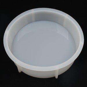 May include: A clear, round silicone mold with a shallow, bowl-like shape. The mold has a smooth, glossy interior and a slightly textured exterior. The sides are straight and the base is curved. The mold is designed for crafting and casting projects.