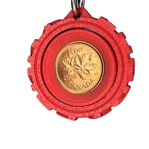 Könnte beinhalten: Ein roter, kreisförmiger Schlüsselanhänger mit einer kanadischen 1-Cent-Münze von 2008. Die Münze zeigt ein Ahornblatt und das Wort „CANADA“. Der Anhänger hat einen strukturierten, zahnradartigen Rand und eine kleine Schlaufe.