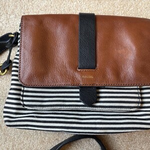 May include: A Fossil crossbody bag featuring a brown leather flap with a vertical black leather strap and a black and white striped canvas body. A gold-tone metal key charm hangs from the black leather shoulder strap on the left side.