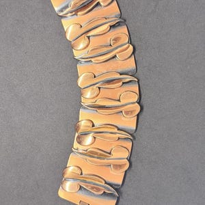 May include: A copper-toned metal link bracelet featuring a series of rectangular segments. Each segment is adorned with a raised, stylized leaf-like motif in a polished copper finish, contrasted against a darkened, oxidized background.