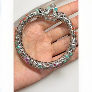 May include: A silver-toned hinged bangle bracelet featuring an intricate openwork metal design. The bracelet is embellished with numerous small, round, multi-colored cabochon stones in shades of red, turquoise, purple, and green set throughout.