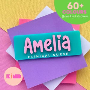 May include: A rectangular teal name badge featuring the name Amelia in raised pink lettering with a small heart above the letter i. Below the name, the text CLINICAL NURSE is printed in white. The badge is layered over several colourful paper squares.