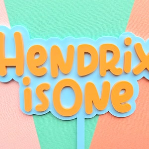 May include: A blue and orange cake topper with the text "Hendrix is One." The text is in orange, with a blue outline, and is set against a pastel background of pink, green, and orange.