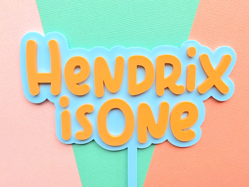 May include: A blue and orange cake topper with the text "Hendrix is One." The text is in orange, with a blue outline, and is set against a pastel background of pink, green, and orange.