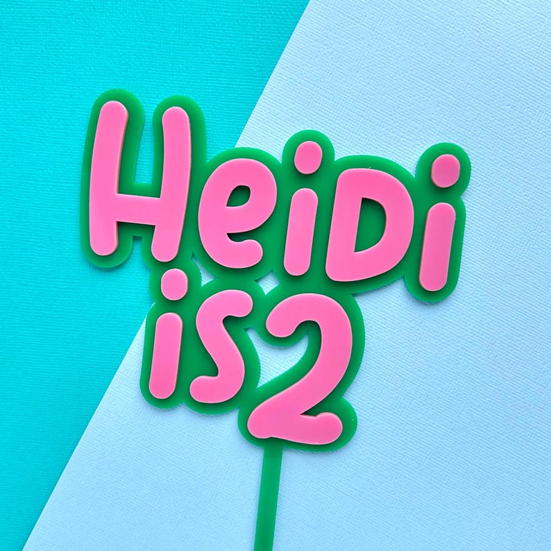 May include: A cake topper with the text "Heidi is 2" in pink letters outlined in green. The topper is on a green stick, set against a turquoise and light blue background. A festive decoration for a second birthday.