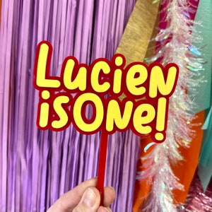 May include: A red and yellow cake topper with the text "Lucien is One!" The text is in a playful, cartoon-like font. The topper is held by a red stick, perfect for a first birthday celebration.