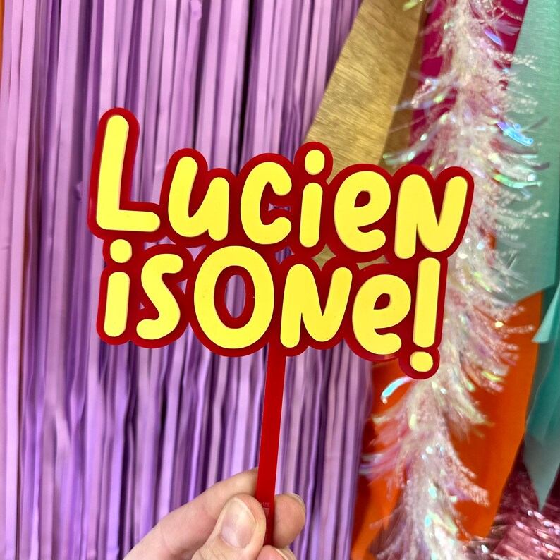 May include: A red and yellow cake topper with the text "Lucien is One!" The text is in a playful, cartoon-like font. The topper is held by a red stick, perfect for a first birthday celebration.