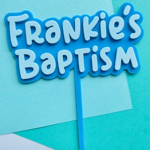 May include: A light blue cake topper with the words "Frankie's Baptism" in white, layered over a light blue base. The topper has a thin, light blue stem for insertion into a cake. The background is a mix of light blue and white paper.