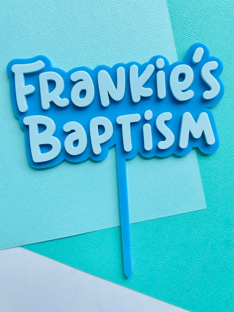 May include: A light blue cake topper with the words "Frankie's Baptism" in white, layered over a light blue base. The topper has a thin, light blue stem for insertion into a cake. The background is a mix of light blue and white paper.