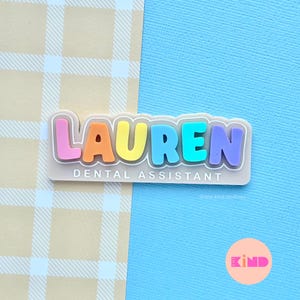 May include: A name tag with the name "LAUREN" in rainbow-coloured letters, outlined in white, and the text "DENTAL ASSISTANT" below. The background is a light tan plaid pattern and a light blue textured surface.