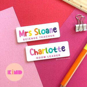 May include: Two white name badges with rainbow text. One says "Mrs Sloane SCIENCE TEACHER" and the other "Charlotte ROOM LEADER". The background is pink and red with a ruler and a binder clip.