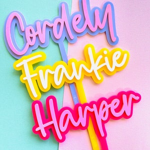 May include: Three colourful name toppers in a script font. The names are Cordely, Frankie, and Harper. The Cordely topper is pink and blue, Frankie is yellow and white, and Harper is pink. Each topper has a stick.