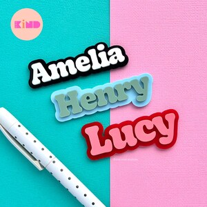 May include: Three name tags with the names "Amelia", "Henry", and "Lucy" in a retro font. The "Amelia" tag is black with white lettering, "Henry" is green with a light blue background, and "Lucy" is pink with a red outline. The background is split between teal and pink.