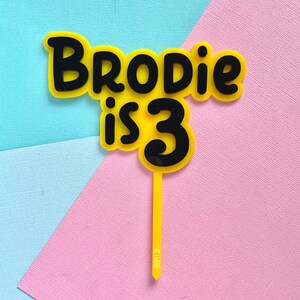 May include: A yellow and black cake topper with the text "Brodie is 3". The text is in a playful font, with a yellow background and black outline. The topper is on a yellow stick, perfect for a birthday cake.