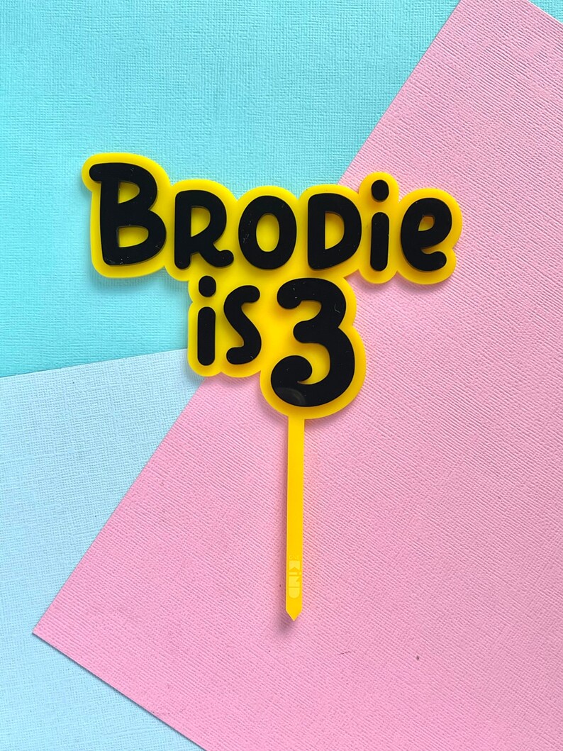 May include: A yellow and black cake topper with the text "Brodie is 3". The text is in a playful font, with a yellow background and black outline. The topper is on a yellow stick, perfect for a birthday cake.