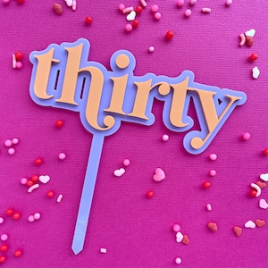 May include: A cake topper with the word "thirty" in orange layered over a lavender background. The topper is on a lavender stick. The background is pink, with scattered sprinkles and heart-shaped candies in various colours.