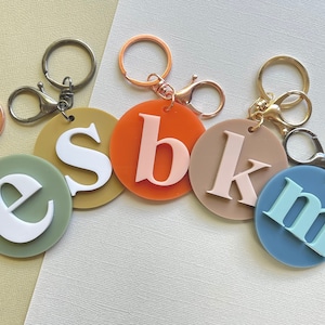May include: A selection of personalised keyrings with circular pendants in various colours. Each keyring displays a different letter in a contrasting colour, attached to a metal ring and clasp. The colour scheme includes green, orange, tan, and blue.