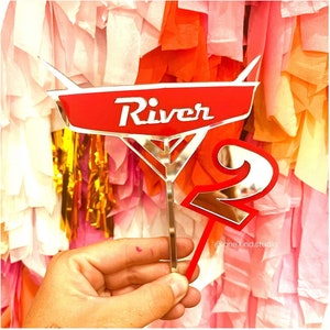 May include: A red and silver cake topper with the name "River" and the number "2". The topper features a car-like design with a silver outline and a red background. The number "2" is also red with a silver outline.