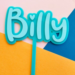May include: A light blue acrylic cake topper with the name "Billy" in a layered, bubble letter style. The topper has a stem for insertion into a cake. The background features yellow, white, and blue geometric shapes.