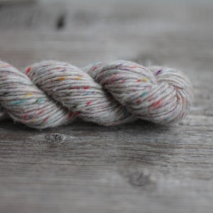 Peut inclure: Gros plan sur une pelote de laine. La laine est de couleur gris clair avec des mouchetures rouges, bleues, jaunes et violettes. La laine est torsadée et semble douce. Le fond est une surface en bois grise.