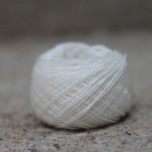 May include: A small, round ball of white yarn featuring a tightly wound, multi-ply construction. The fibers are smooth and uniform, creating a consistent texture across the entire surface of the spherical object, which is presented in a clear focus.