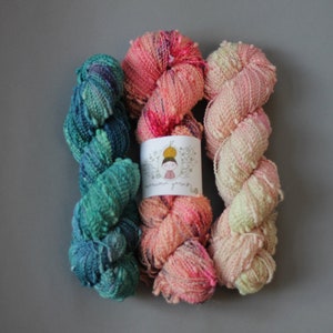 Puede incluir: Tres ovillos de hilo en diferentes colores. Los ovillos son rosa, verde y melocotón. El hilo es texturizado y tiene un aspecto peludo. Los ovillos están etiquetados como "remarkable yarns".