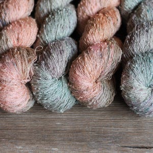 May include: Four skeins of textured yarn arranged in a row. The yarn features a variegated color palette of soft peach, muted sage green, and light gray tones. Each skein has a nubby, irregular surface texture with small flecks of darker color.