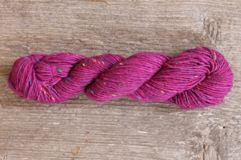 Puede incluir: Un ovillo de hilo magenta con motas de fibras multicolores. El hilo est&aacute; retorcido y dispuesto en una bobina suelta.