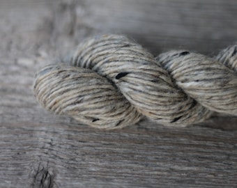 May include: A close-up of a skein of yarn in a light gray and brown color with black flecks. The yarn is soft and fluffy, and it is perfect for knitting or crocheting.