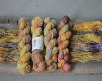 May include: Five skeins of hand-dyed yarn in shades of yellow, orange, and purple. The yarn is arranged in a row on a wooden surface. The yarn is labeled "Happy Yarns".