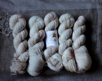 May include: Six skeins of off-white yarn with subtle brown and purple variegation, arranged on a textured gray surface. A small label with a whimsical illustration is visible. A crocheted doily is in the upper left corner.