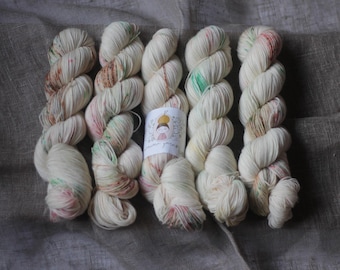 May include: Five skeins of off-white yarn with subtle speckles of pink, green, and brown. A label with a drawing of a person is visible. The yarn is arranged on a neutral-colored fabric, creating a soft, inviting texture.