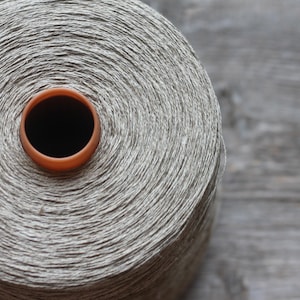 Puede incluir: Una gran bobina cilíndrica de hilo gris claro texturizado, enrollado firmemente alrededor de un núcleo central. La bobina presenta un anillo central de plástico liso de color marrón rojizo en la parte superior, contrastando con el hilo neutro.