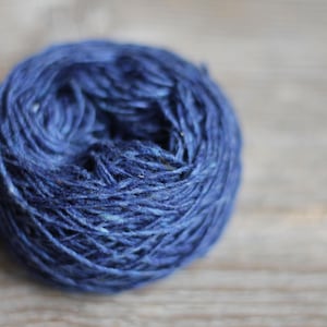 Puede incluir: Una madeja de hilo azul índigo, bien enrollada, descansa sobre una superficie de madera marrón claro. El hilo tiene una apariencia texturizada, lo que sugiere una fibra natural. Ideal para proyectos de punto o ganchillo.