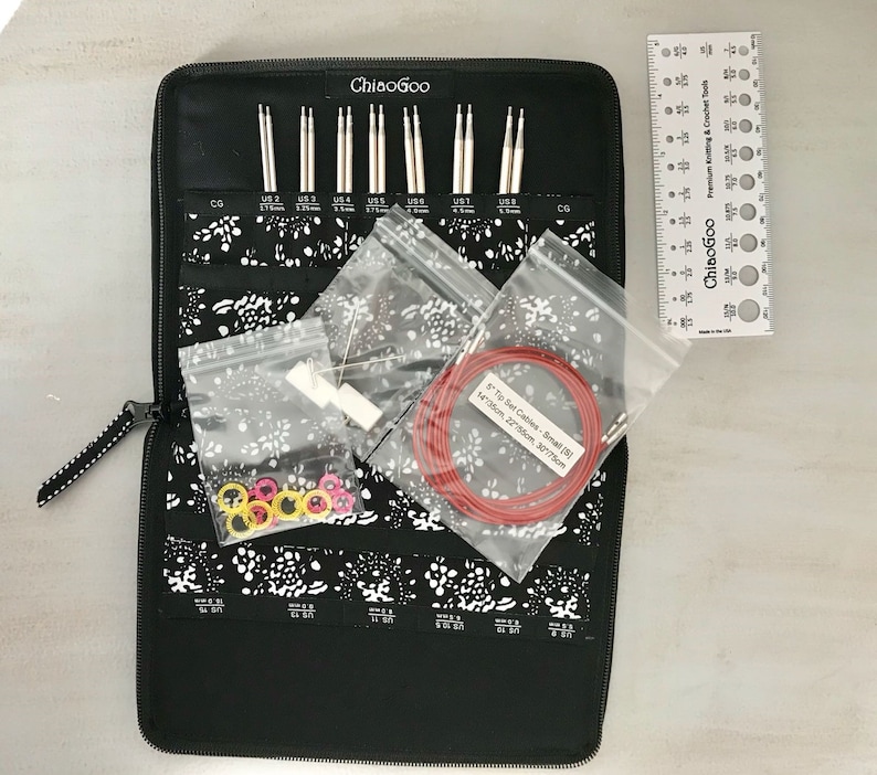 May include: A black zippered case with a white and black floral pattern holds a set of ChiaoGoo knitting needles. The needles are organized by size and include a variety of sizes. The case also includes a ruler, a set of stitch markers, and a set of cables.