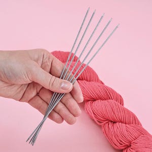 Op de afbeelding: Een hand houdt een bundel zilveren breinaalden vast, boven een streng koraalkleurig garen. De naalden zijn dun en puntig, en het garen is zacht en getextureerd. De achtergrond is effen roze.